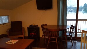 A cozy living room with a fireplace, a television mounted on the wall, and a view of a snowy landscape through a large window. - Cute Beachfront Guest Unit on Lake (Saugatuck)
