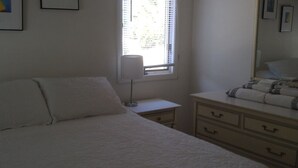 A bedroom with a bed, a bedside table with a lamp, a dresser, and a window with blinds. - Cute Beachfront Guest Unit on Lake (Saugatuck)