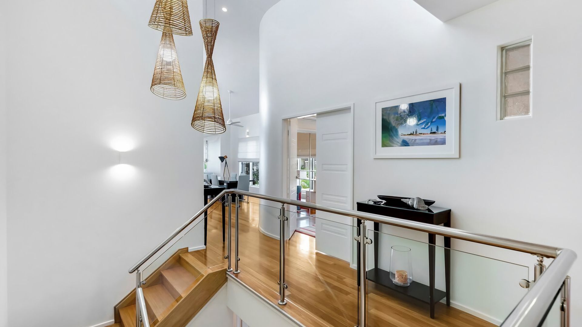 Un interior moderno con una escalera de madera, barandilla de cristal y vistas a un comedor.