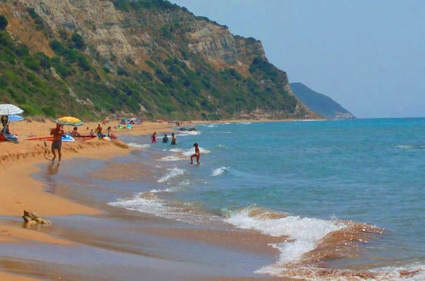 Une plage où l’on voit des gens se baigner et se promener le long du rivage, une falaise en arrière-plan et un ciel dégagé.
