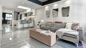 Ein modernes Wohnzimmer mit einem grauen Sofa, einem rosa Hocker und einem Couchtisch. An der Wand hängen drei Spiegel, im Hintergrund ist eine Küchenzeile zu sehen, und ein großes Fenster sorgt für viel Licht.