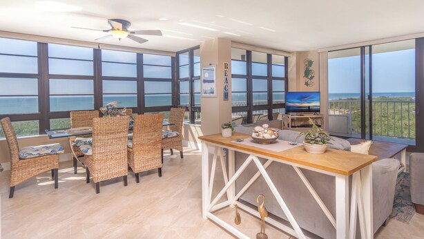 A spacious room with large windows, wicker furniture, a wooden table, and a television.