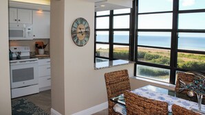 A kitchen with white cabinets, a microwave, and an oven, leading to a dining area with wicker chairs and a view of the ocean.