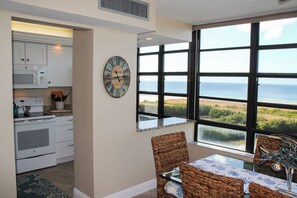 A kitchen with white cabinets, a microwave, and an oven, leading to a dining area with wicker chairs and a view of the ocean.
