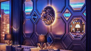 Un espace bar moderne avec un décor mural géométrique, une sculpture représentant une tête de lion et un aménagement avec des tables et des lampes.