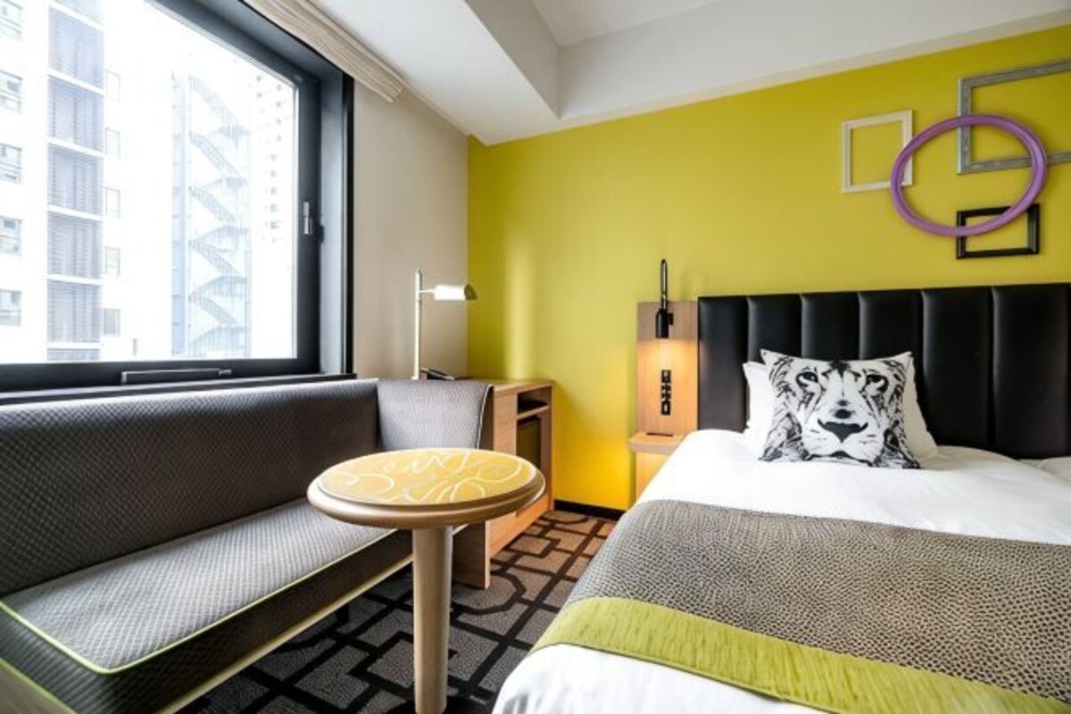Een hotelkamer met een bed, een slaapbank, een klein tafeltje, een nachtkastje en een raam met uitzicht op gebouwen.