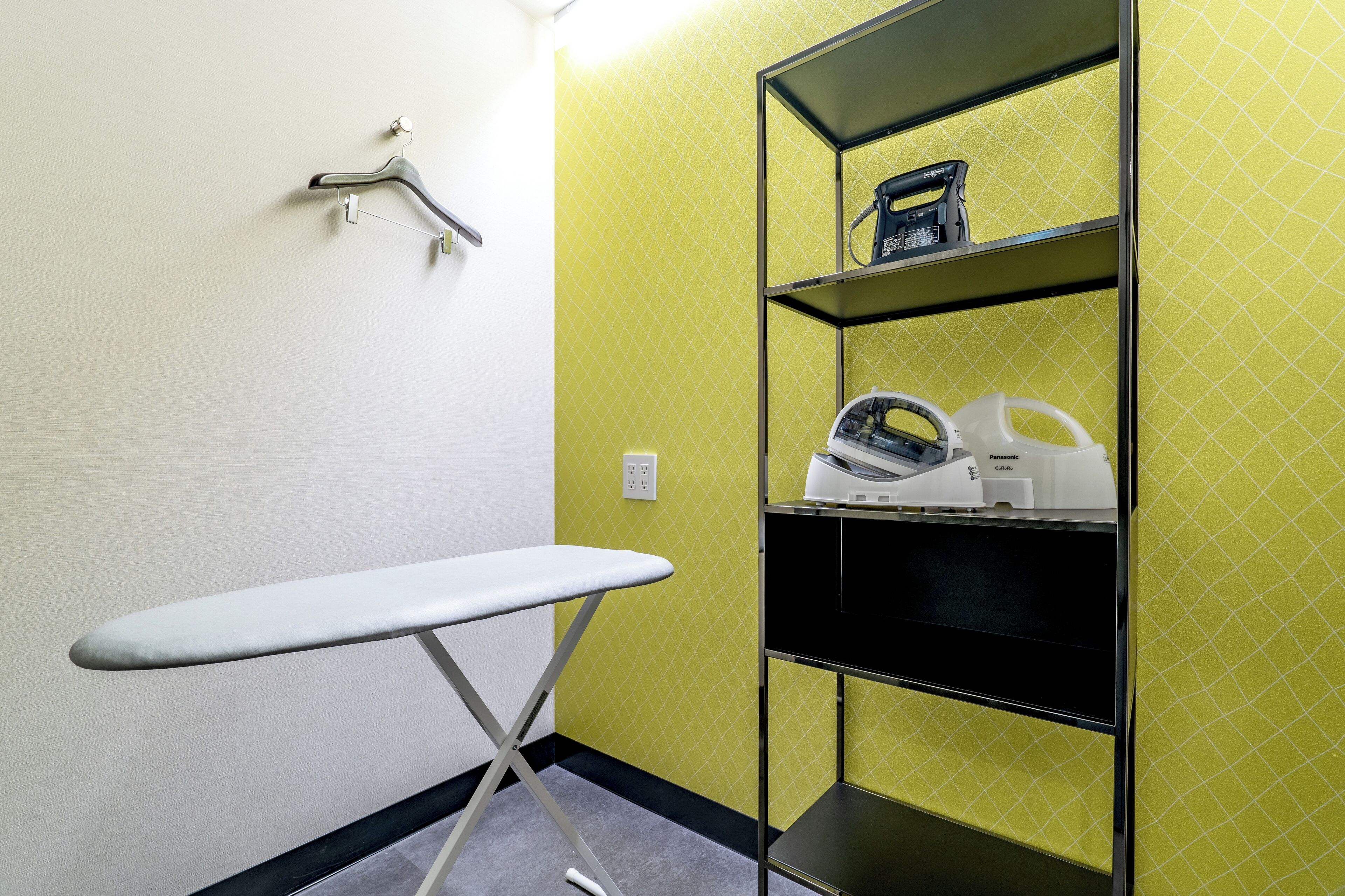 A room with a white ironing board, a black and white heater, and a shelf with a kettle and a Panasonic coffee maker.