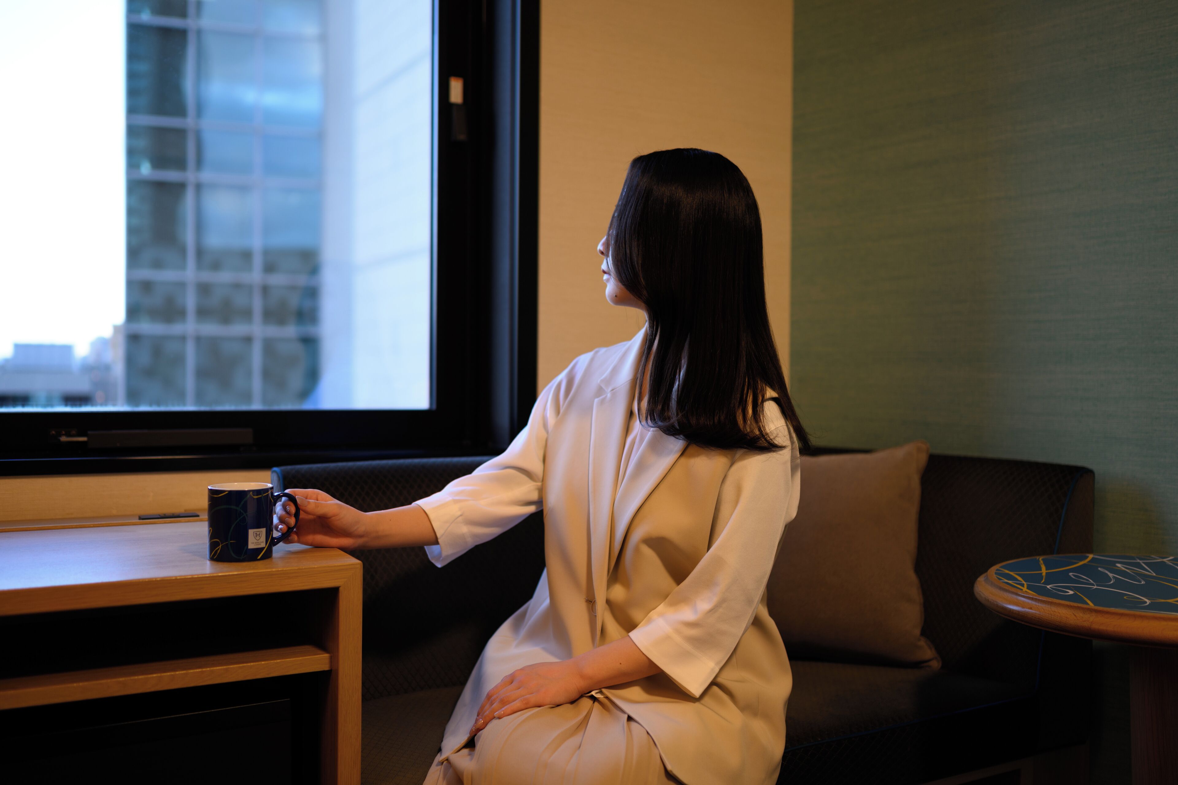 A woman sitting on a couch with a coffee mug on a side table, facing a large window with a cityscape view.