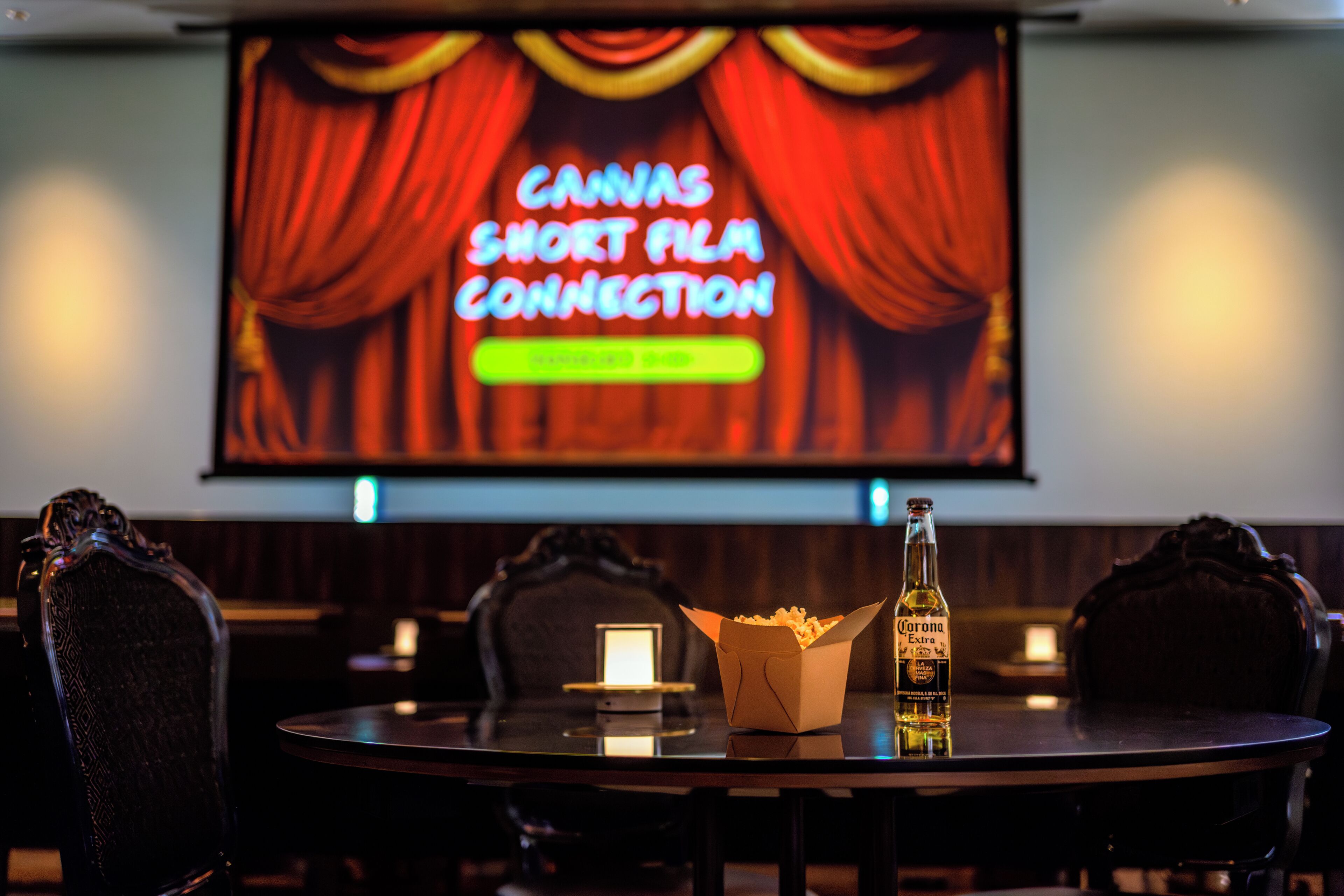Una sala de cine con una pantalla que muestra 'CANVAS SHORT FILM CONNECTION' y una mesa con palomitas de maíz y una botella de cerveza Corona.