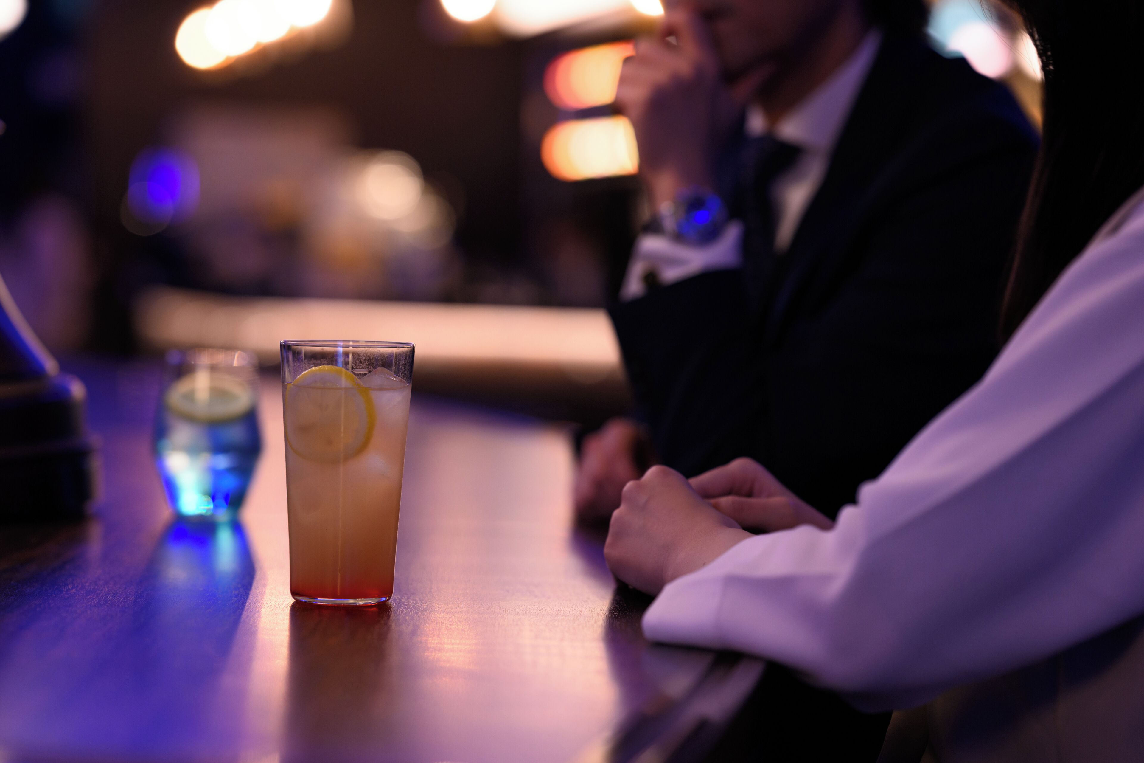 A bar setting with a glass of drink and a person seated at the bar.