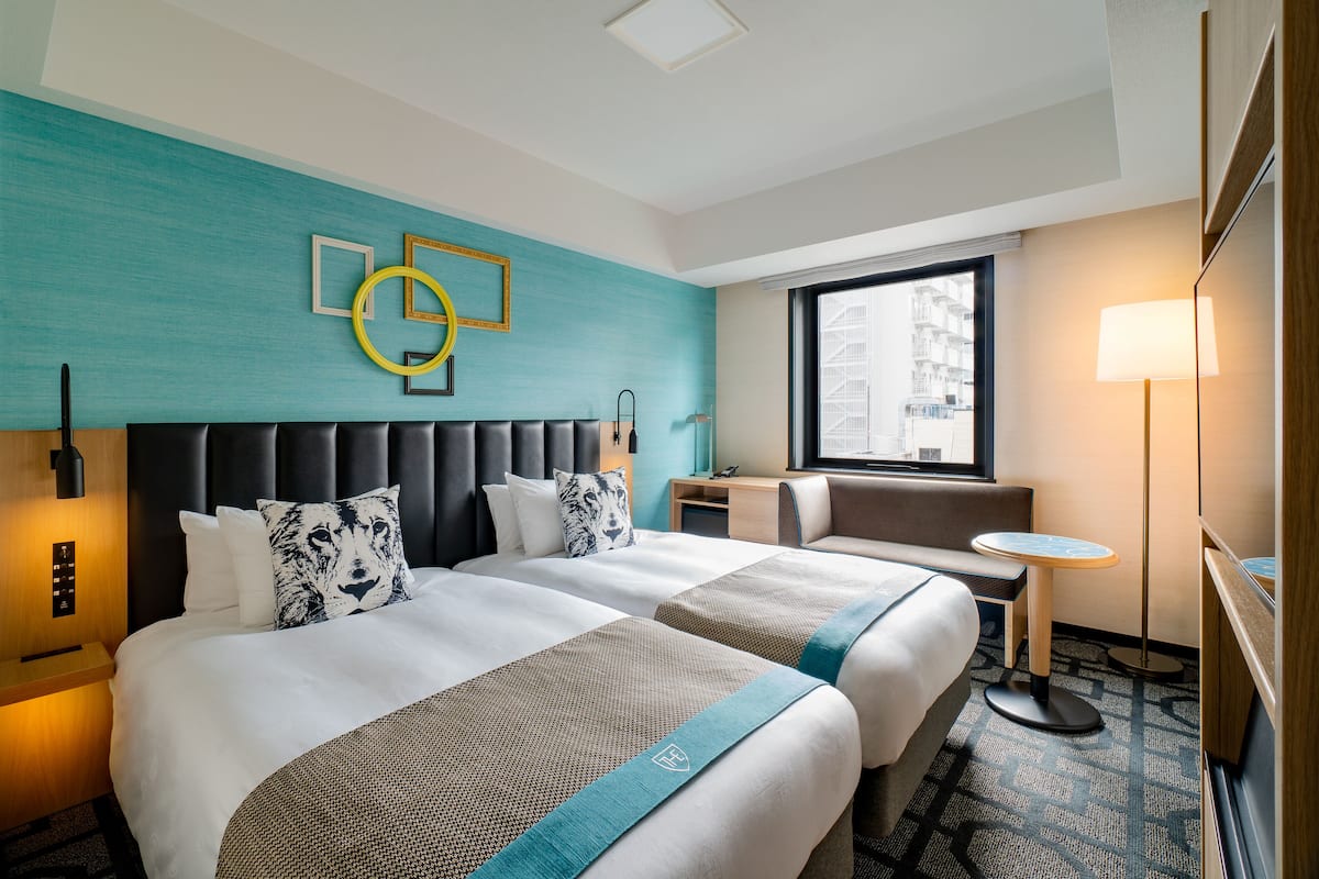 Une chambre d’hôtel avec deux lits, une table ronde, un banc, une lampe et une fenêtre donnant sur la ville.
