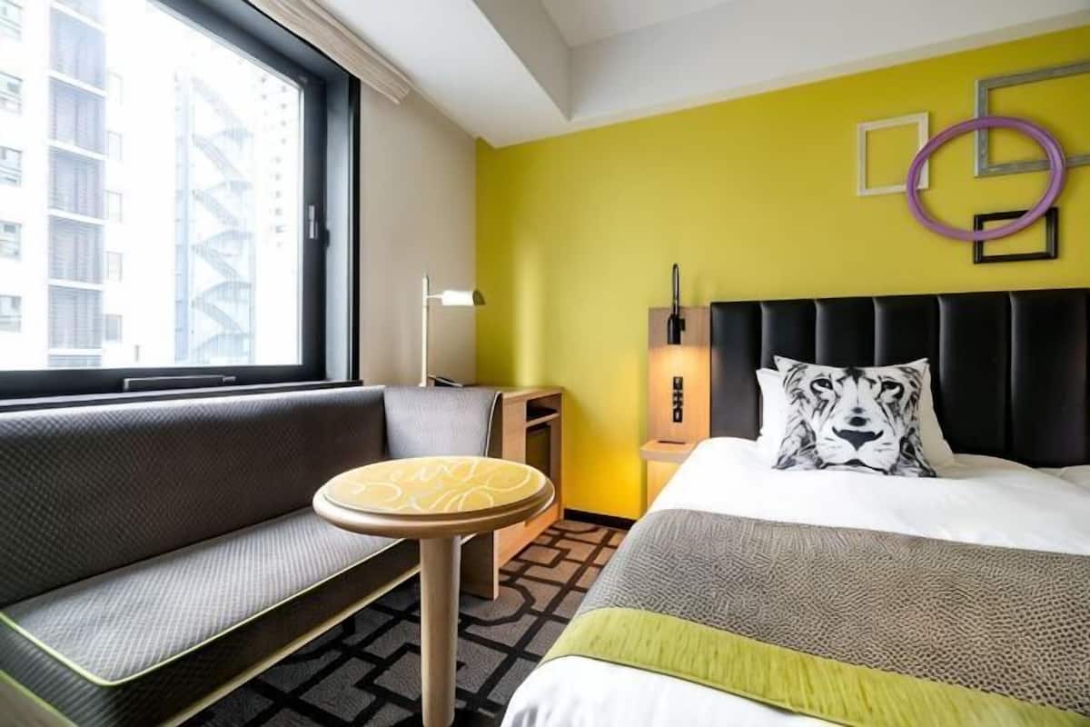 Une chambre d’hôtel avec un lit, un canapé, une petite table, une table de chevet et une fenêtre donnant sur des immeubles.