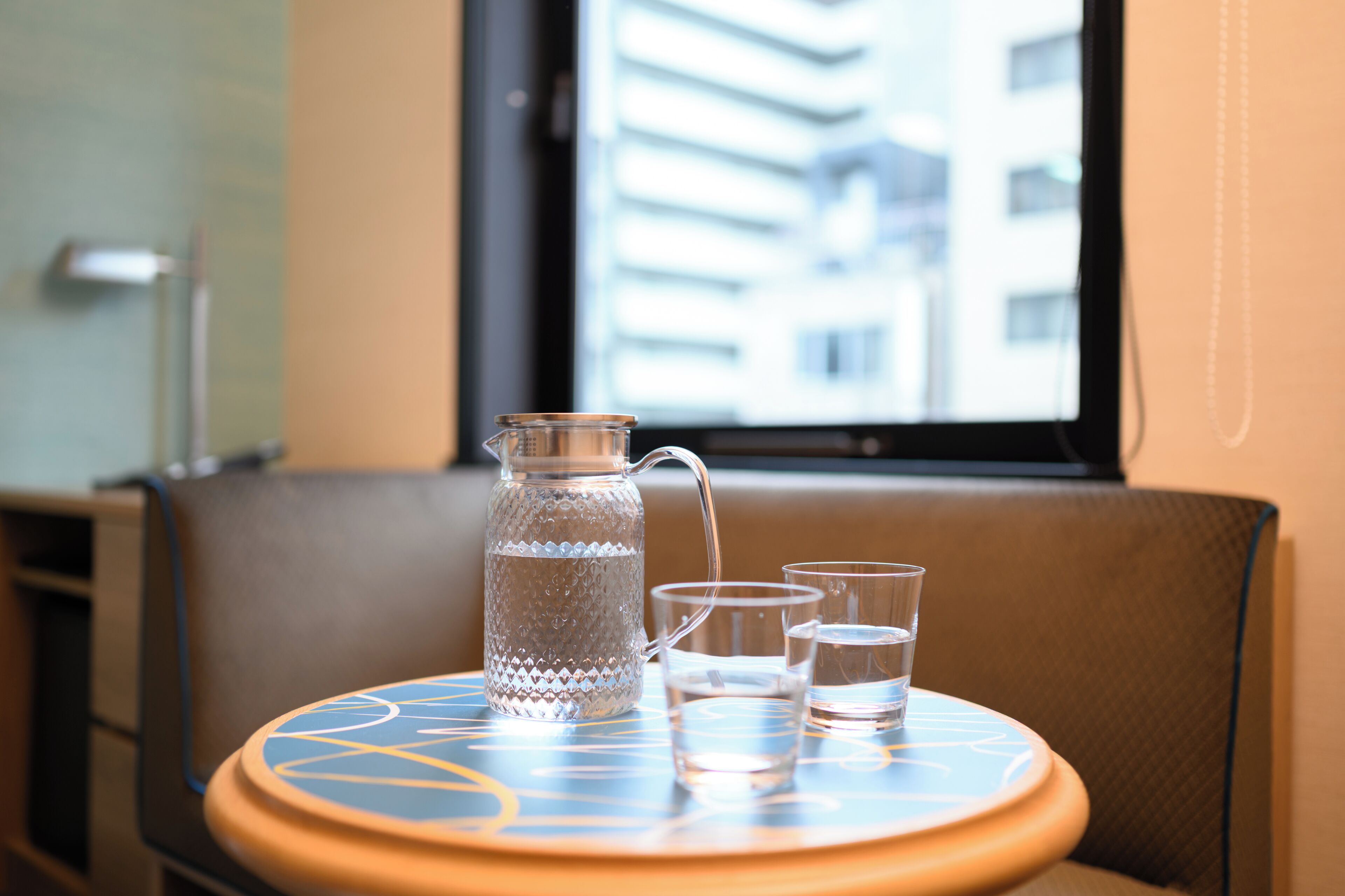 A table with a water pitcher and glasses, a window with a view of buildings, and a chair.