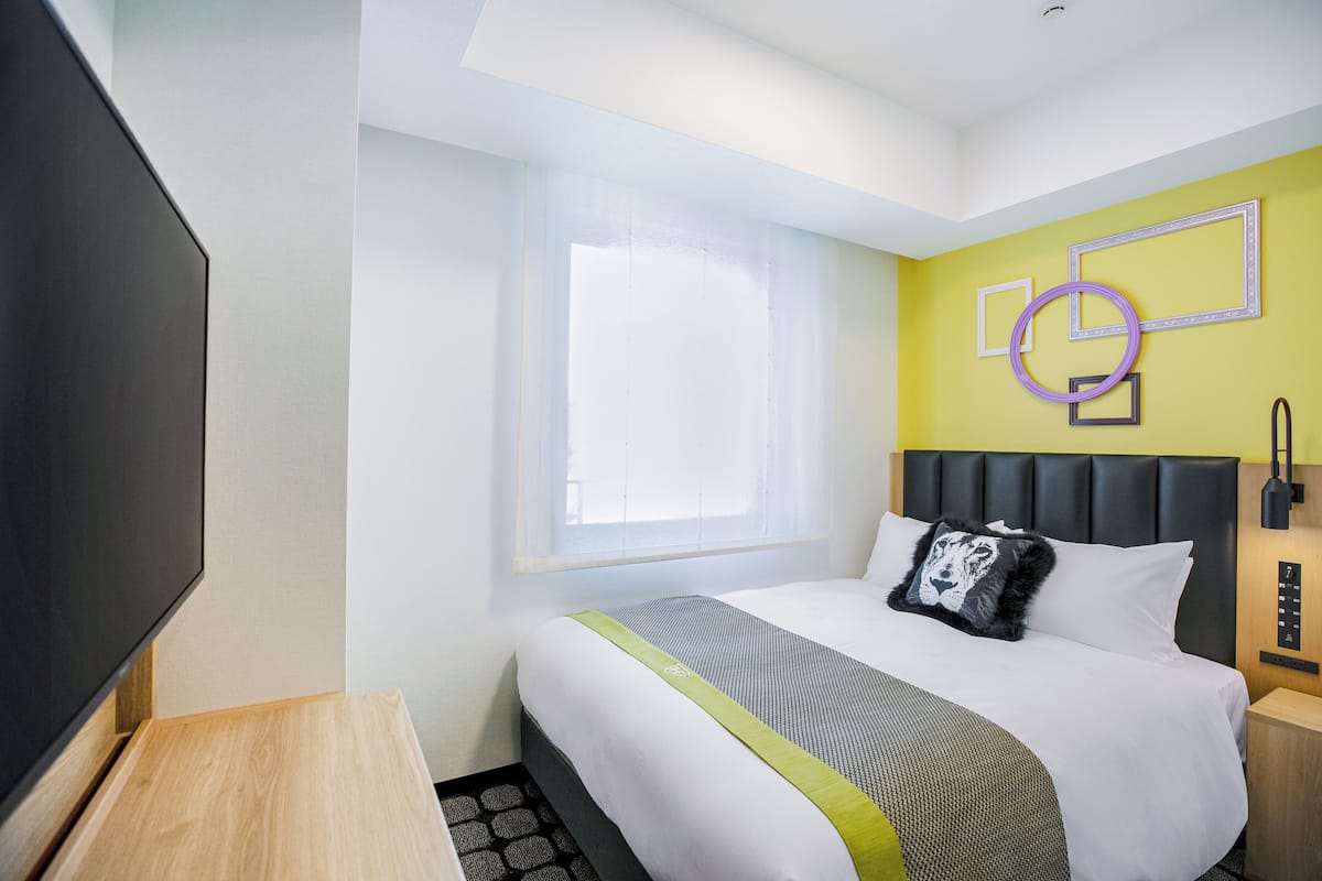 Une chambre d’hôtel moderne dotée d’un grand lit, d’un téléviseur à écran plat, d’une fenêtre avec stores et d’un mur orné de motifs géométriques.