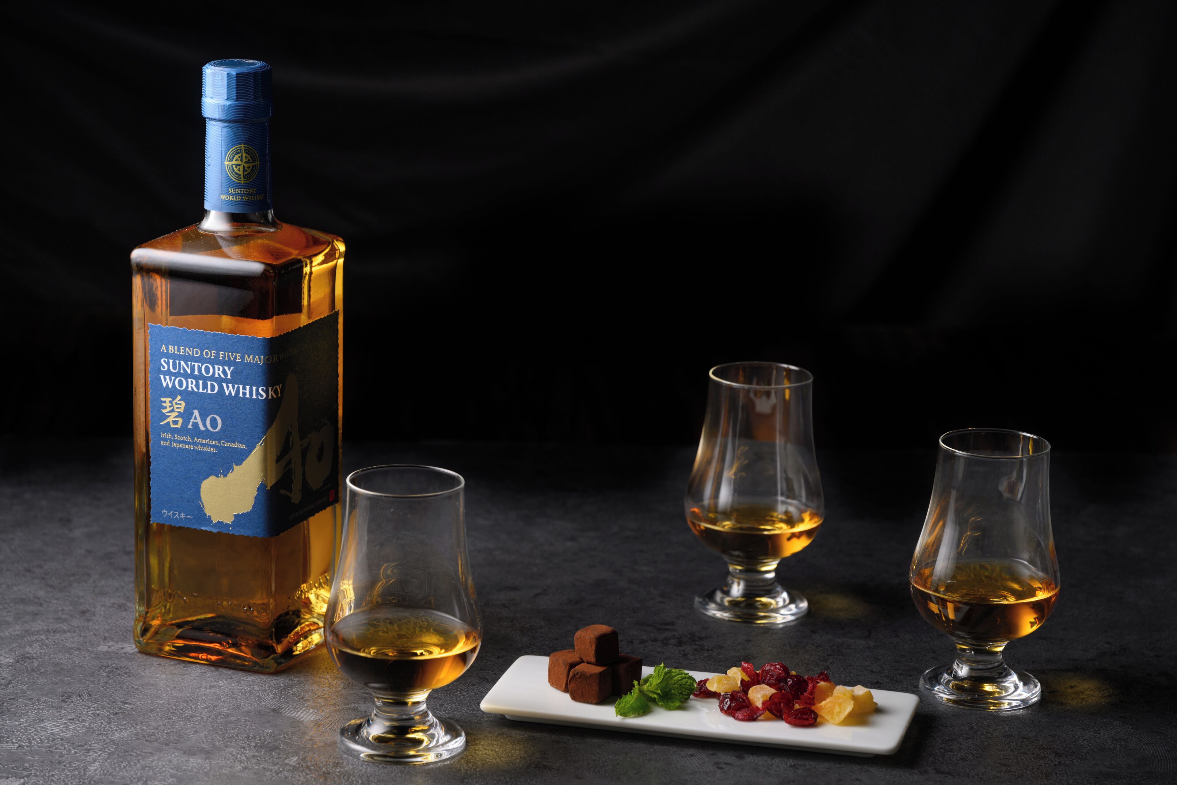 A bottle of Suntory World Whisky AO, two glasses, and a plate with assorted snacks.