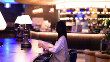 A woman sitting at a bar with a drink, a lamp, and a bar counter in a dimly lit room with ambient lighting.