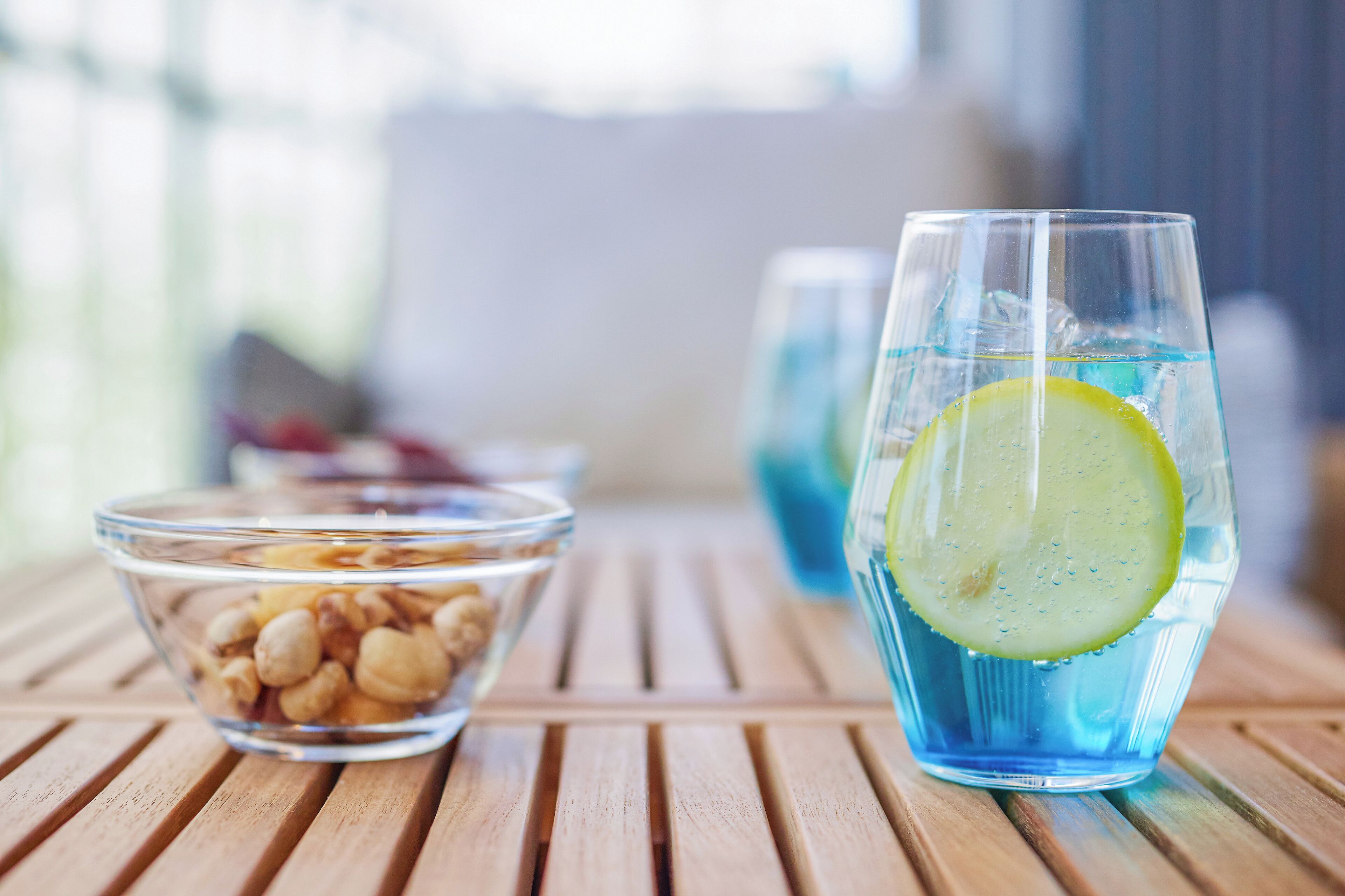 A glass of blue beverage with a lemon slice, a bowl of nuts, and a clear glass container on a wooden table.