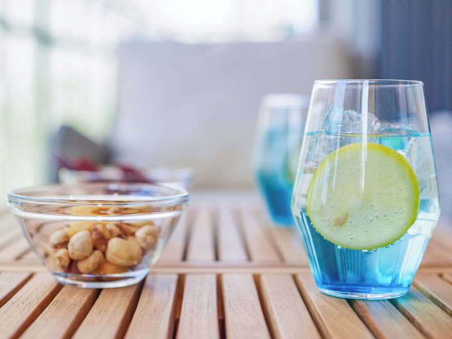 A glass of blue beverage with a lemon slice, a bowl of nuts, and a clear glass container on a wooden table.