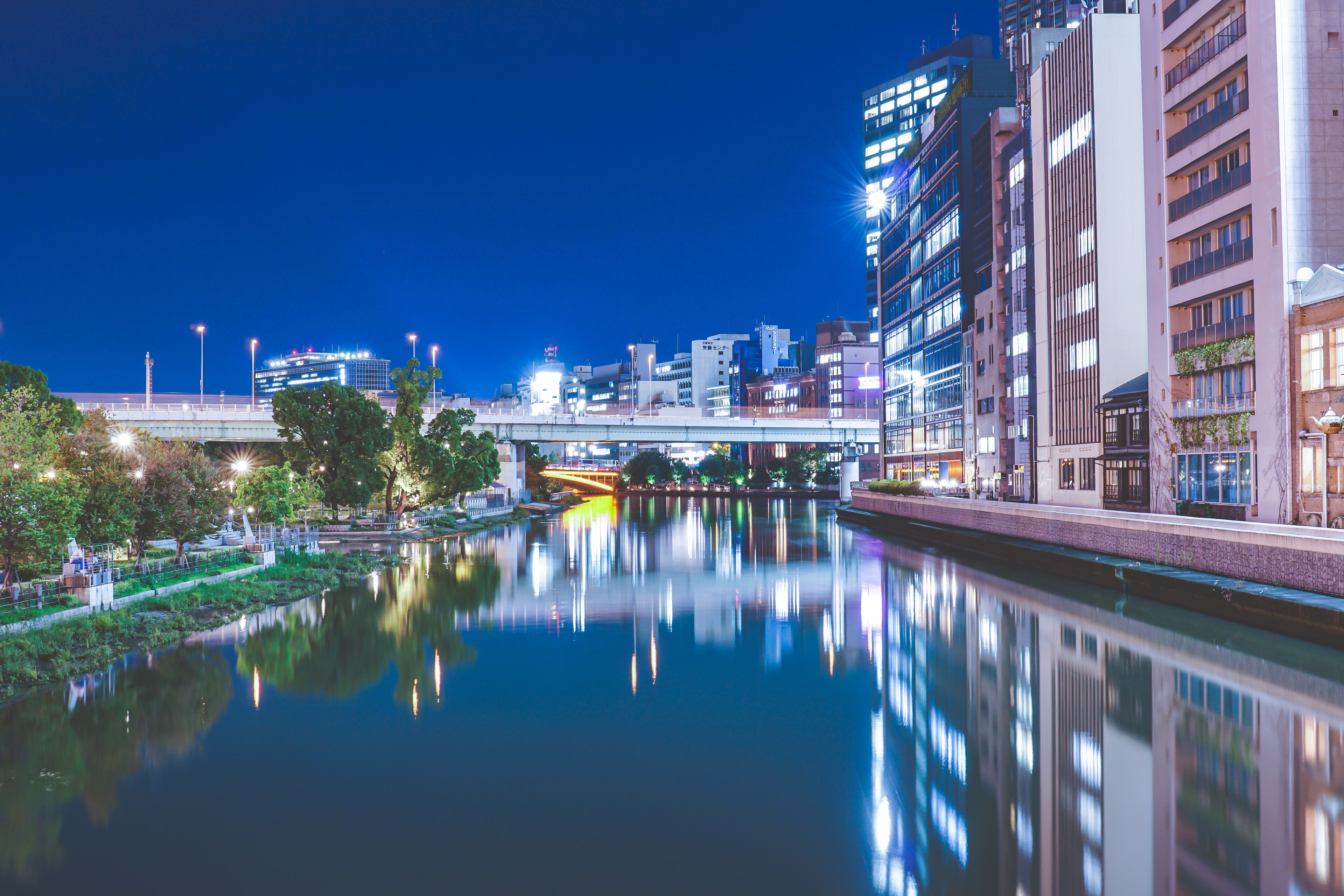 A nighttime cityscape with a river reflecting buildings and a bridge.