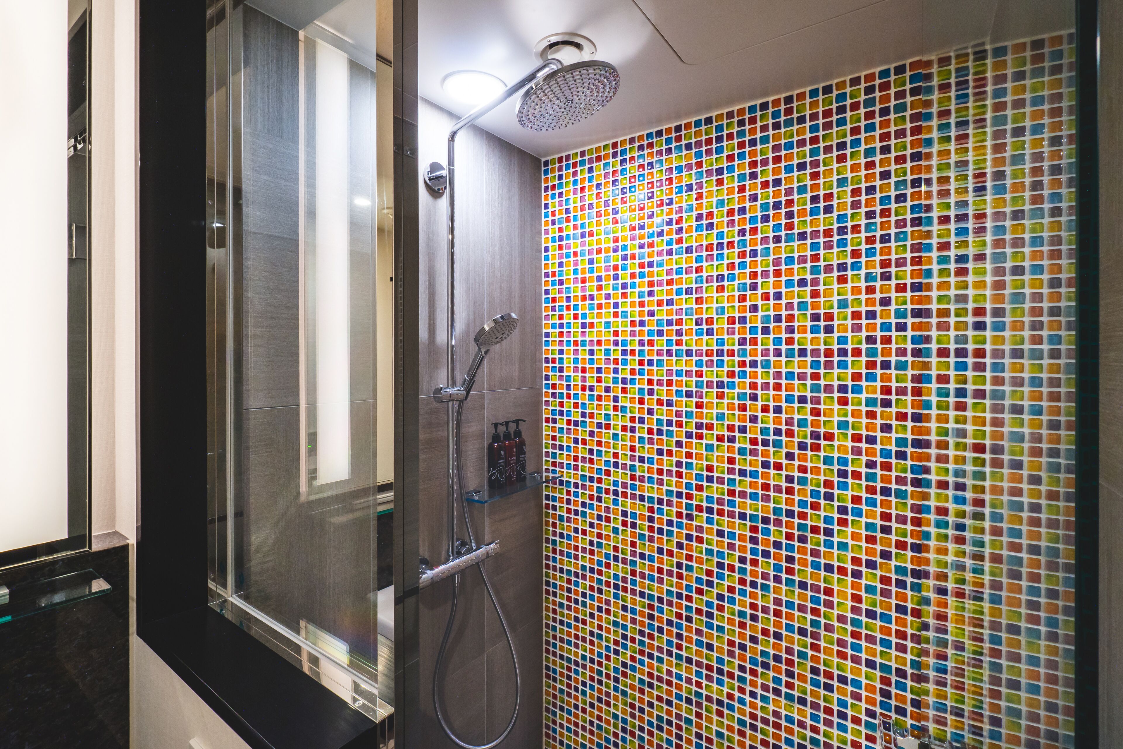 A modern bathroom with a glass shower enclosure, a handheld showerhead, and a colorful mosaic tile wall.