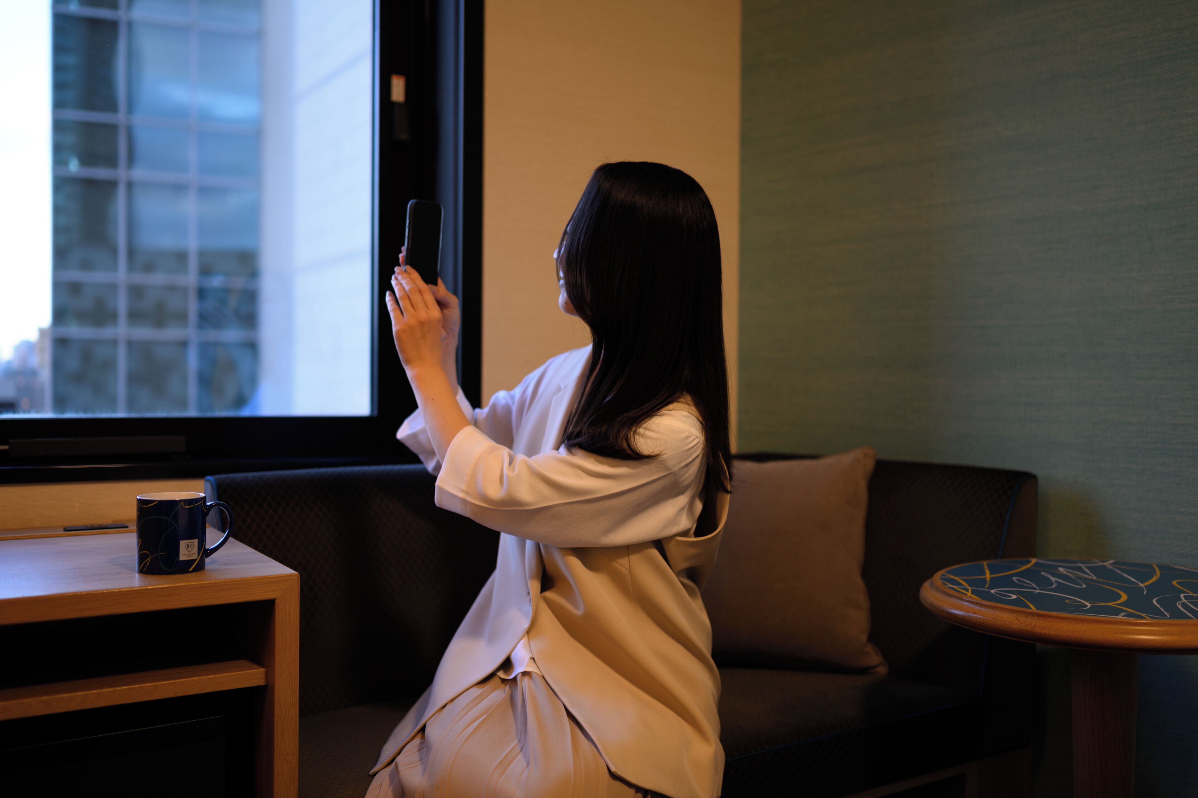 Une personne est assise sur un canapé, un ordinateur portable est posé sur ses genoux, une tasse de café se trouve sur une petite table à côté, et une fenêtre offre une vue sur des immeubles.