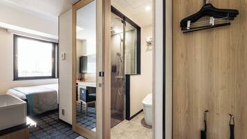 A compact hotel room with a bed, a small desk, a shower area, and a wooden wardrobe.