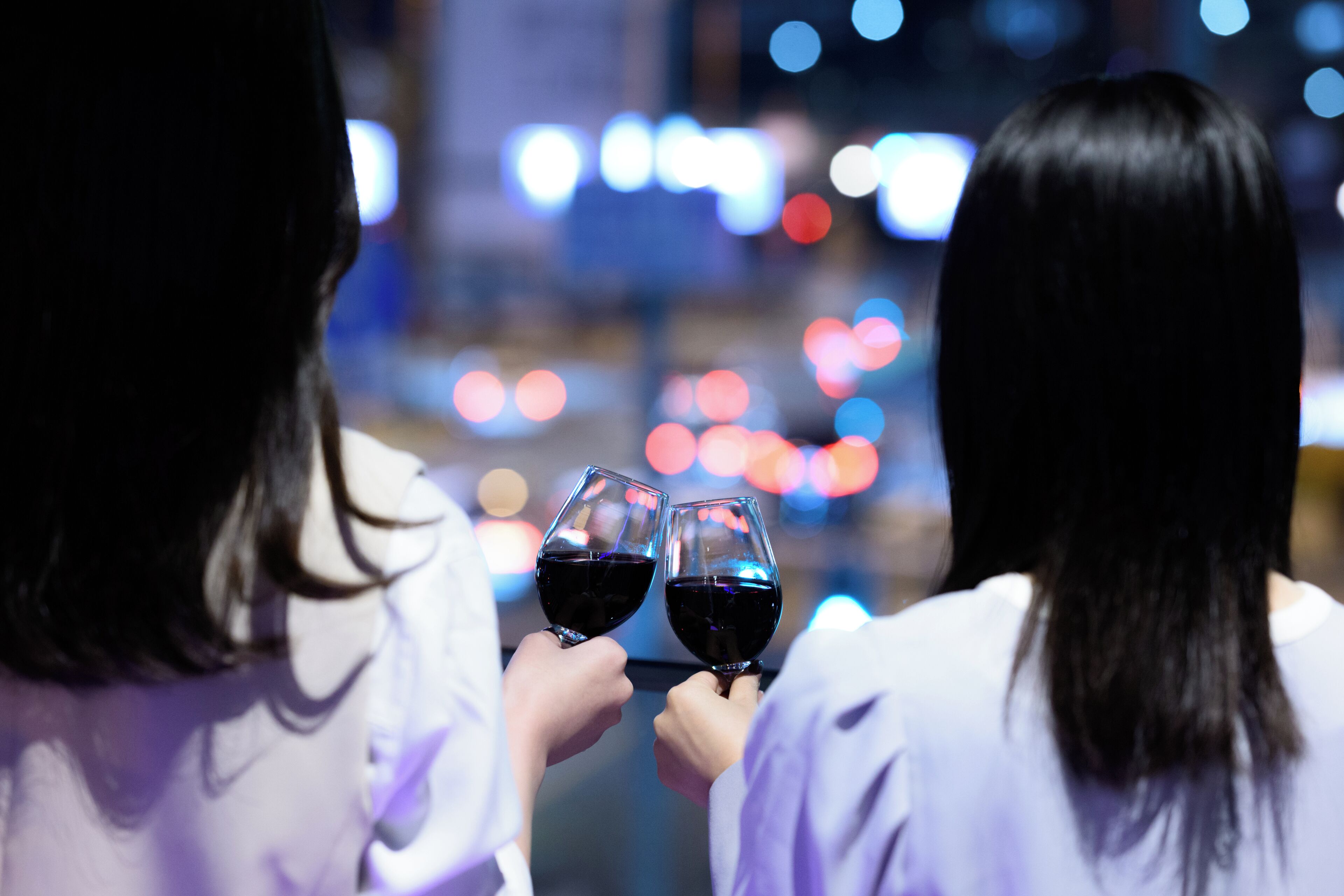 Two people holding wine glasses, facing away from the camera, with a cityscape in the background.