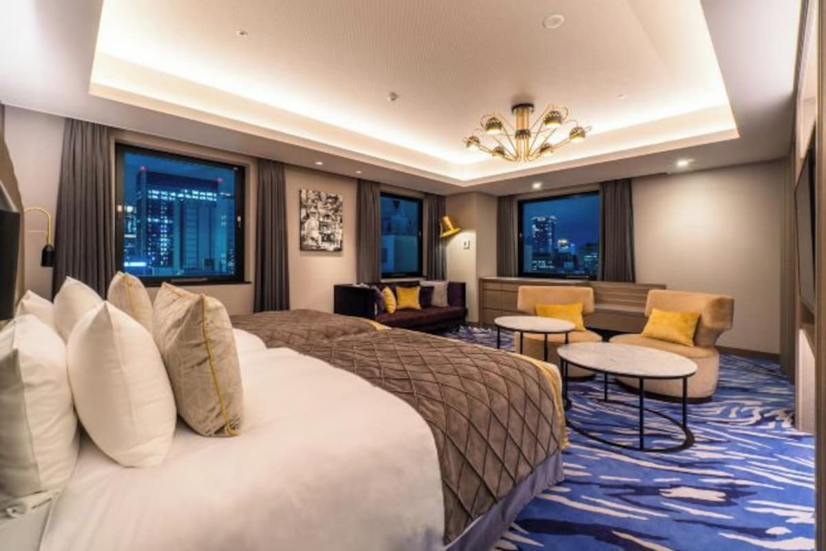 Habitación de hotel con una cama grande, un sofá, dos sillones, una mesita y vistas a la ciudad a través de amplios ventanales.