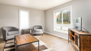 Een moderne woonkamer met twee fauteuils, een salontafel, een televisie op een houten kast en een groot raam met uitzicht op bomen.