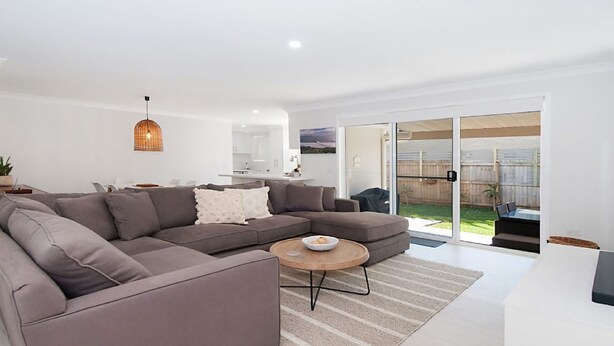 A modern living room with a sectional sofa, a round coffee table, and a sliding glass door leading to a backyard. - Kay's Hideaway (Lennox Head)