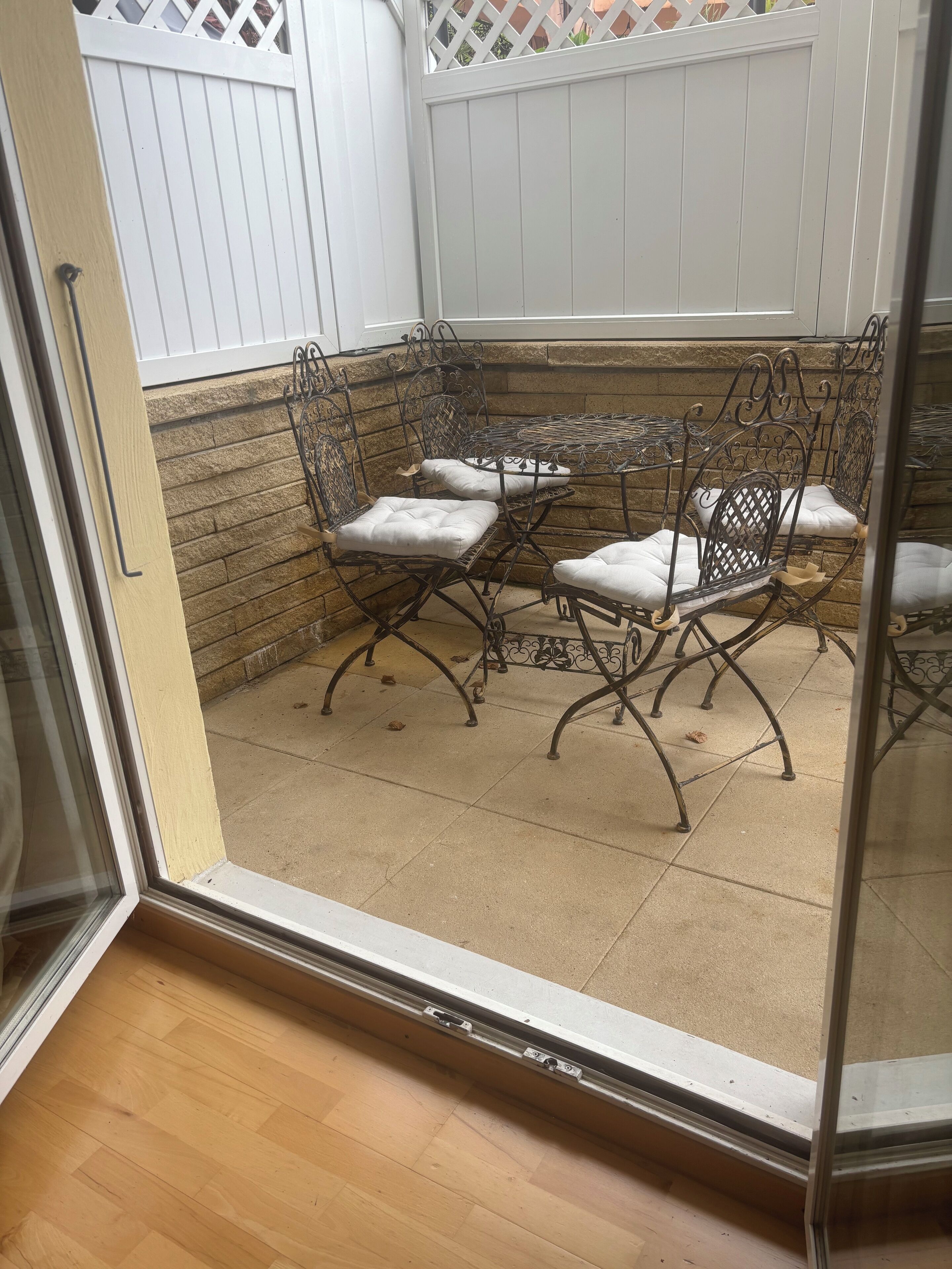 Un patio con una mesa metálica y cuatro sillas, una cerca blanca y un muro de piedra.