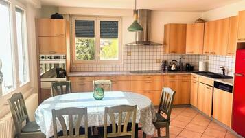 Une cuisine avec des meubles de rangement en bois, une table à manger avec des chaises, un réfrigérateur rouge et une fenêtre avec des stores.