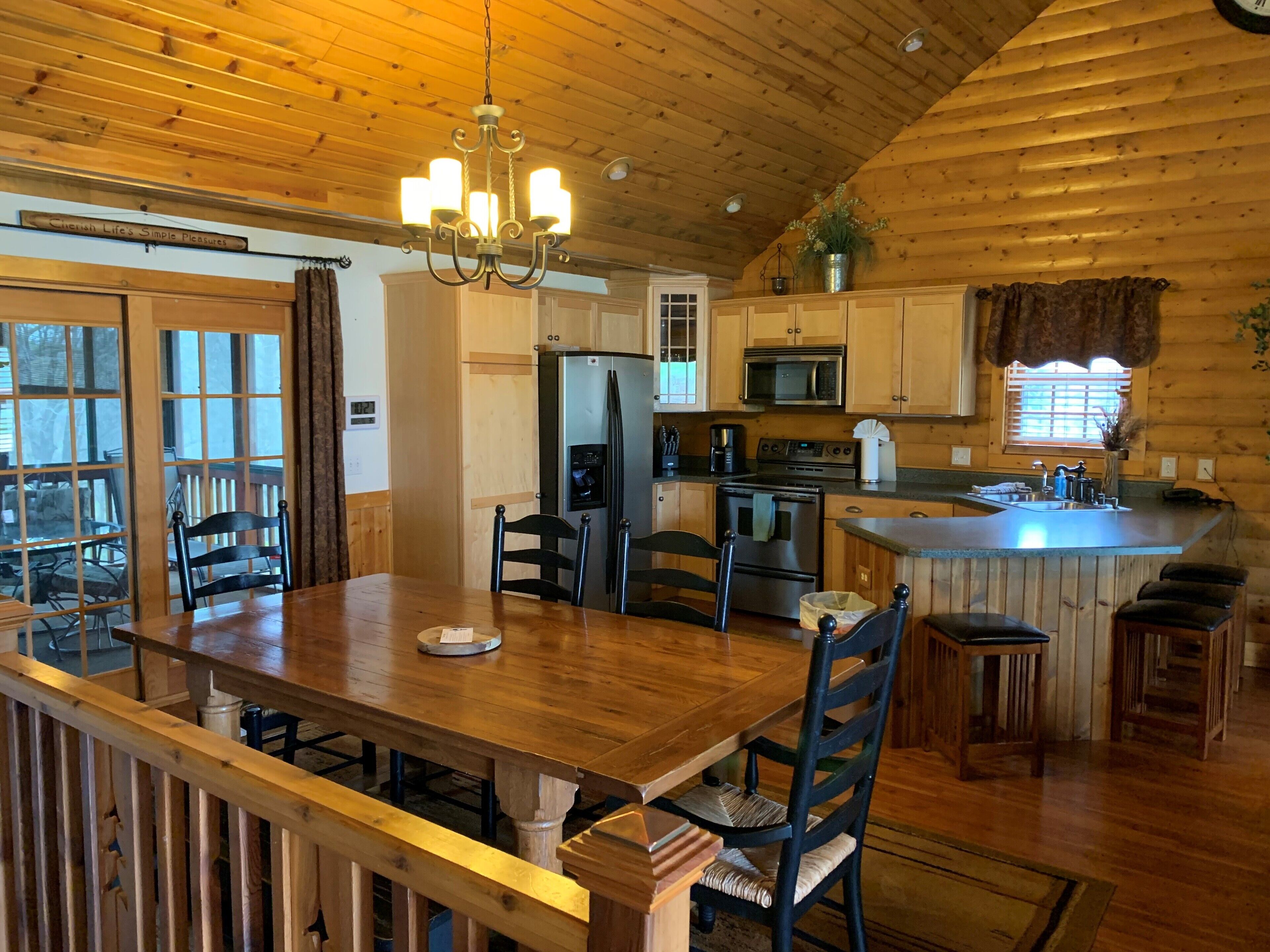 A wooden cabin interior with a dining area, kitchen, and a large window.