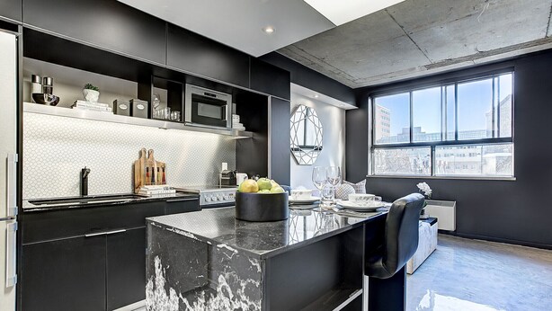 A modern kitchen with a black countertop, stainless steel appliances, and a large window providing a view of the city. - Roomy Loft near Guy-Concordia Metro (Montreal)