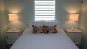 A bedroom with a bed, two bedside tables, a window with blinds, and two lamps. - Beautiful Garden Pool, Walk to Town   (Vieques)