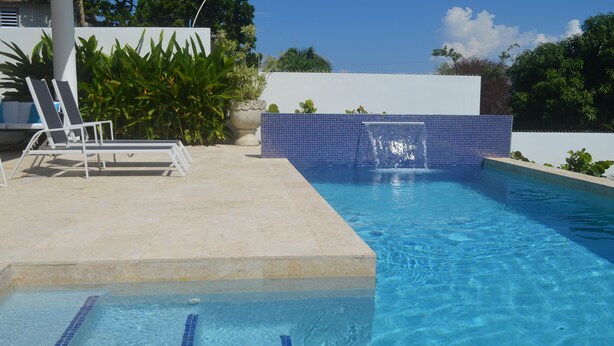 A swimming pool with clear blue water, a tiled wall, and a white lounge chair. - Beautiful Garden Pool, Walk to Town   (Vieques)