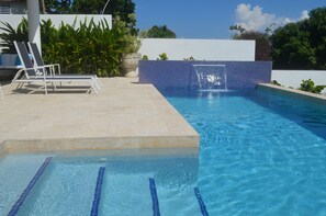 Una piscina amb aigua blava cristal·lina, una paret de rajola i una cadira de relaxació blanca.