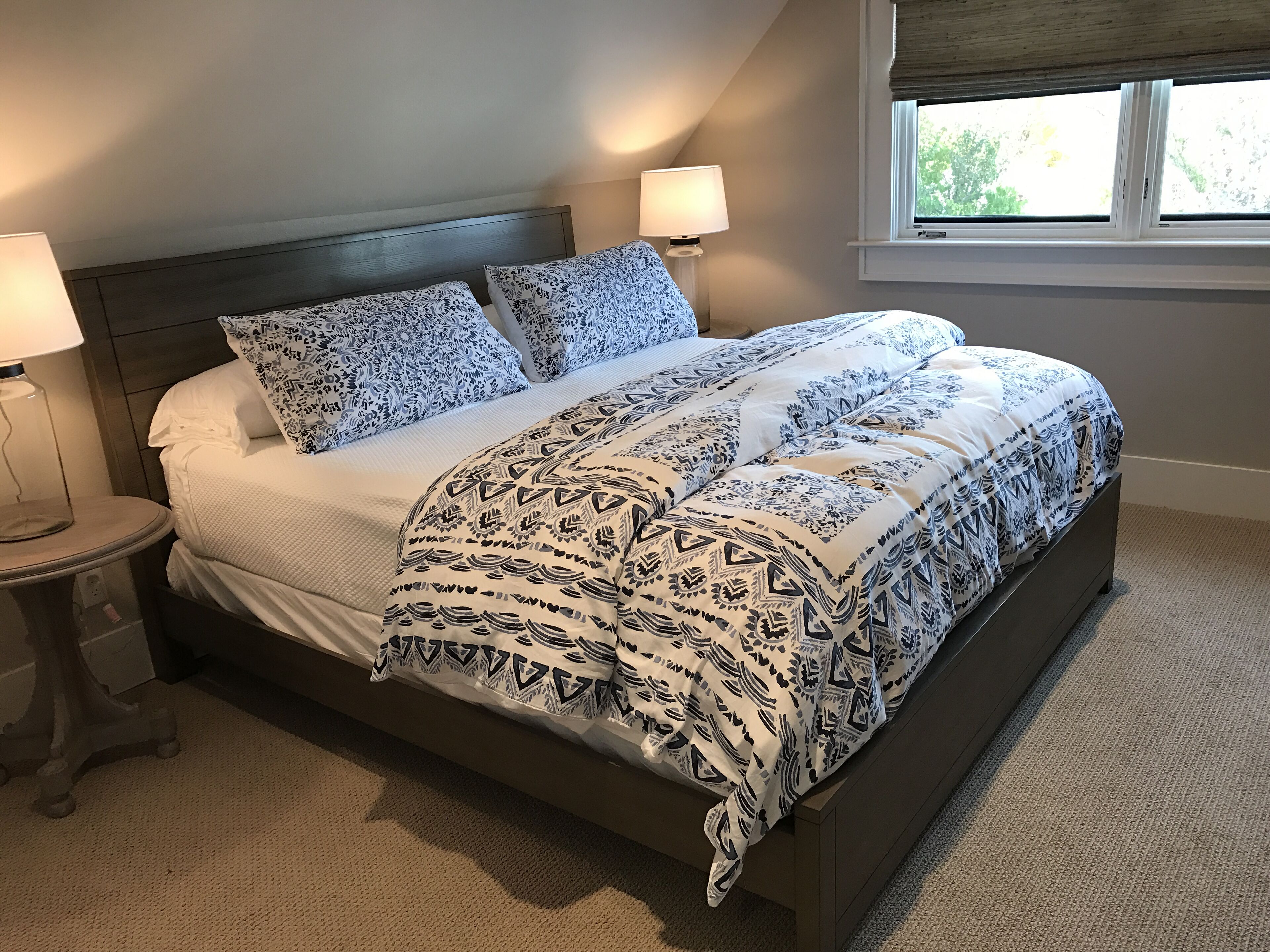 Una cama bien hecha con ropa de cama estampada, una lámpara de noche y una ventana con vistas a zonas verdes.