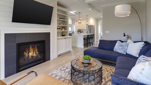 A modern living room with a fireplace, a large flat-screen TV, a blue sofa, and a dining area in the background.