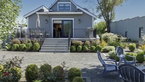 A two-story house with a front porch, a staircase, and a garden with trimmed bushes and a few chairs.
