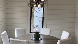 A round dining table with white upholstered chairs, a geometric pendant light, and a glass dome over a green object.