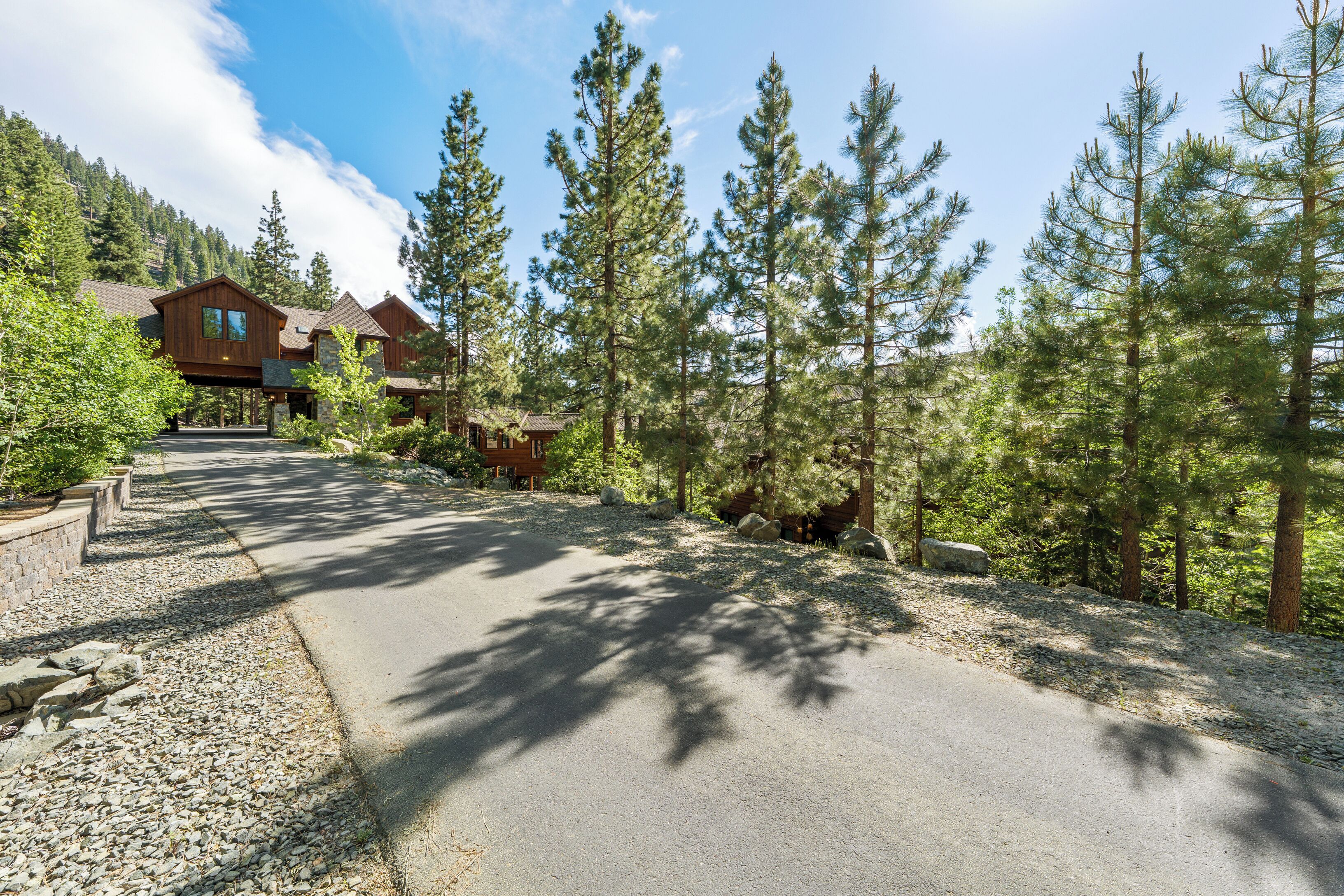 A lodge with a paved driveway, surrounded by tall pine trees and a mountain backdrop.