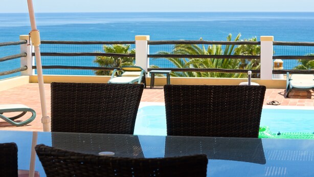 Ein Balkon mit Korbstühlen und einem Glastisch mit Blick auf einen Pool und den Ozean.