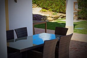 Eine Terrasse im Freien mit einem Tisch aus Glasplatten und Korbstühlen, umgeben von einem angelegten Garten und einer Steinmauer.