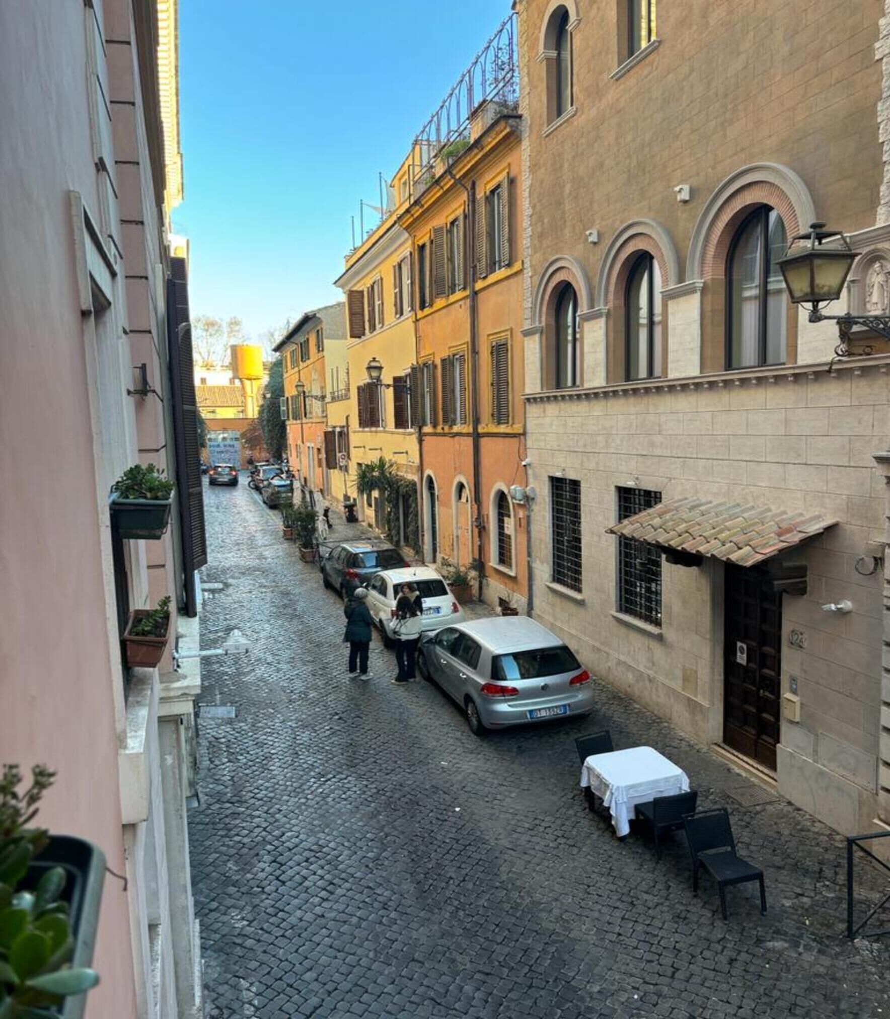 A narrow cobblestone street lined with historic buildings, a parked car, and a table with chairs set up outside a building.