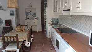Eine Küche mit weißen Schränken, einem Holztisch mit bunten Tischdecken und einer Pendelleuchte, sowie einer gefliesten Rückwand.