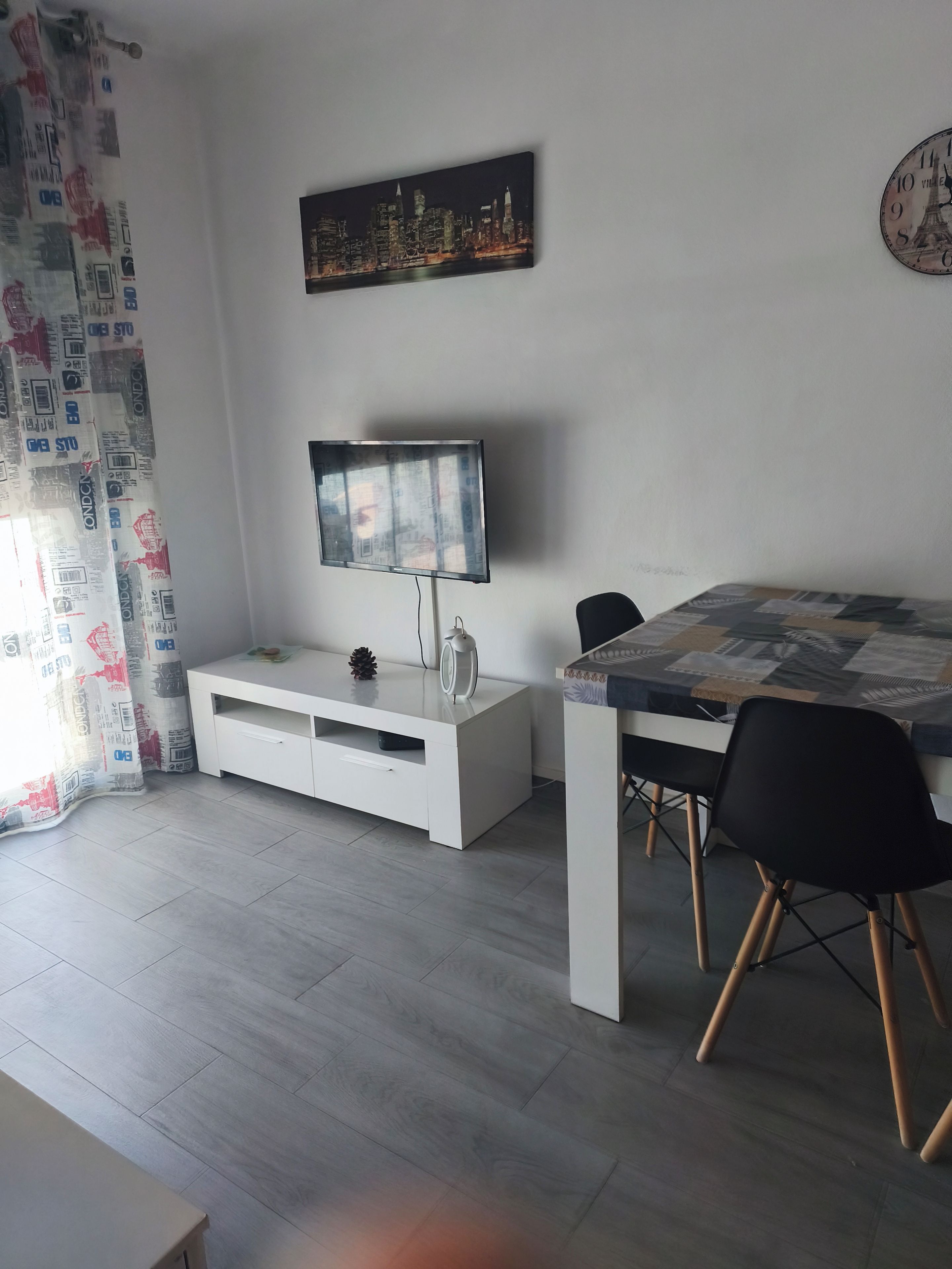 Een moderne woonkamer met een flatscreen televisie, een eettafel met stoelen, een witte tv-kast, en een stadsgezicht schilderij aan de muur.