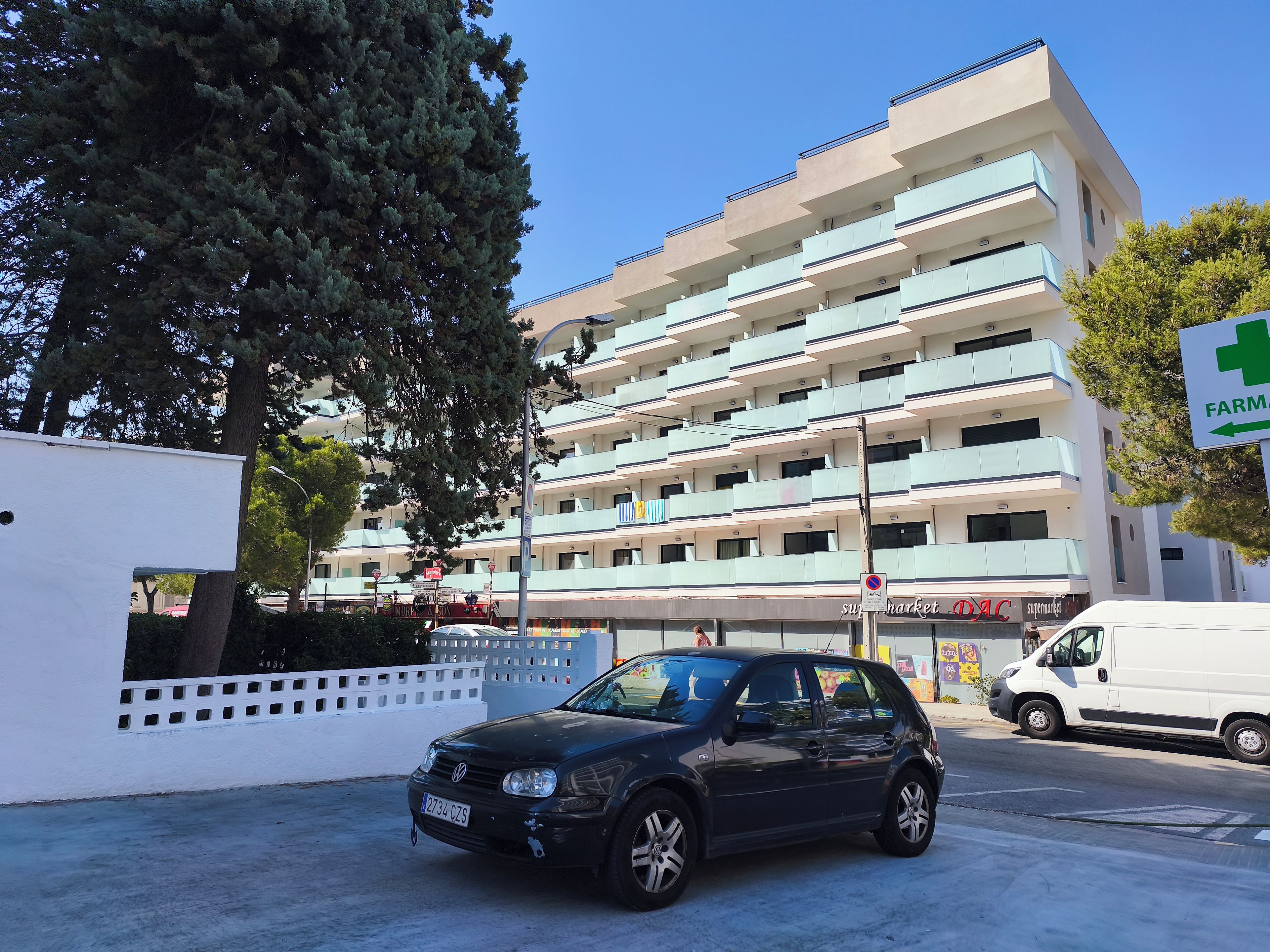 Un edificio de varios pisos con un letrero de farmacia, una camioneta blanca y un coche negro estacionados en la entrada.