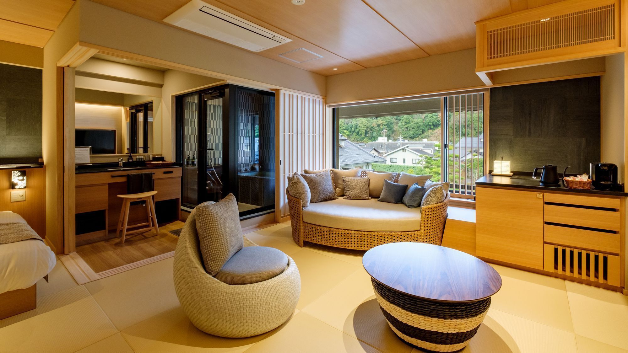 A modern hotel room with a sofa, a round table, a kitchen area, and a view of the outside.
