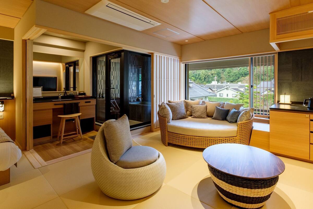 A modern hotel room with a sofa, a round table, a kitchen area, and a view of the outside.